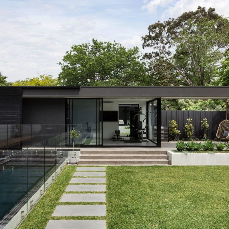 "Modern house exterior with black minimalist design, glass-walled pool, gym area, and landscaped garden